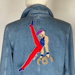 May include: A blue denim jacket with a colorful embroidered patch of a gymnast performing on the rings. The patch features a red leotard, blue and white stripes, and a purple hat.