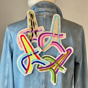 Könnte beinhalten: Eine Jeansjacke mit bunten gestickten Patches in verschiedenen Formen und Designs. Die Patches sind aus Filz und haben verschiedene Farben, darunter Rot, Blau, Grün und Gelb.