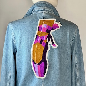 Peut inclure: Veste en jean bleu clair avec un grand écusson brodé représentant une chaussure de danse de ballet. L'écusson présente un motif de couleurs or, violet et rose. La veste a un col classique et une fermeture boutonnée.