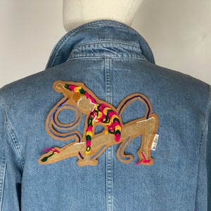 Peut inclure: Une veste en jean bleu avec un patch brodé coloré au dos. Le patch représente une figure stylisée en position de gymnastique, avec un schéma de couleurs bleu, jaune, rose et vert.