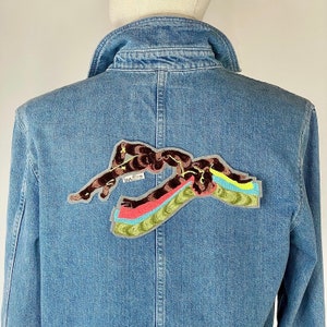 Peut inclure: Une veste en jean avec un patch brodé coloré représentant une personne nageant. Le patch est gris, marron, rose, bleu et jaune. Le texte "budlee" est visible sur le patch.