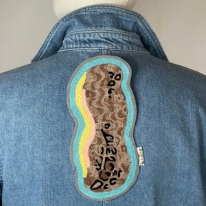 Peut inclure: Une veste en jean bleu avec un patch brodé marron, jaune et rose au dos. Le patch a la forme d'une cacahuète et porte le texte "706..." brodé dessus.