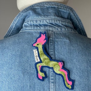 Peut inclure: Une veste en jean avec un patch brodé de cerf vert et rose sur le dos. Le patch porte l'inscription "HUDSON".