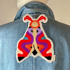 May include: A denim jacket with a colourful embroidered moth patch on the back. The moth is red, orange, purple and blue.