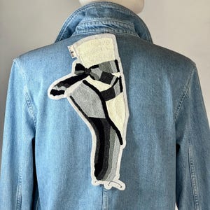 Peut inclure: Veste en jean bleu clair avec un grand patch abstrait dans le dos. Le patch présente un motif dans les tons de noir, de gris et de crème. La veste a un col classique et une fermeture boutonnée.