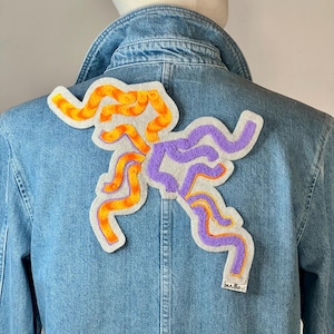May include: A denim jacket with a large, colorful patch sewn on the back. The patch is made of felt and features a design of orange and purple wavy lines.