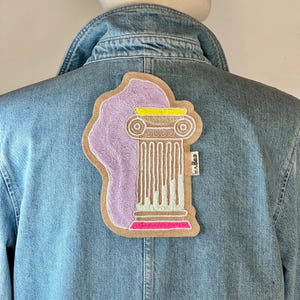 Può includere: Giacca di jeans azzurra con una toppa decorativa con un disegno a colonna. La toppa ha un bordo marrone, una parte superiore di perline lavanda e dettagli gialli e rosa. La giacca ha un colletto classico e bottoni.
