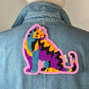 Peut inclure: Veste en jean avec un grand écusson brodé coloré représentant un chat stylisé. L'écusson présente un contour rose et un motif de chat avec des détails bleus, jaunes, violets et noirs.