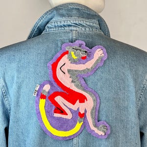 Peut inclure: Veste en jean bleu clair avec un grand écusson brodé coloré. L'écusson représente une panthère stylisée en rose, rouge, gris et jaune, sur fond violet. La veste a un col classique et des fermetures boutonnées.