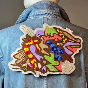 Peut inclure: Veste en jean avec un grand écusson brodé coloré dans le dos. L'écusson présente un motif abstrait avec des formes marron, jaune, rouge, violet, vert et bleu. L'écusson est bordé de crème.