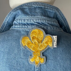 以下が含まれることがあります： 青いデニムジャケットに金色の刺繍が施されたフルール・ド・リスのパッチ。パッチはジャケットの後ろ、襟の近くに縫い付けられています。