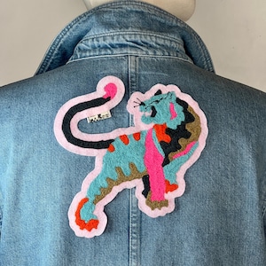 Peut inclure: Veste en jean bleu clair avec un écusson brodé d'un tigre coloré. Le patch représente un tigre turquoise avec des détails roses, oranges et marron, soulignés de rose. Un tourbillon noir et rose est au-dessus du tigre.