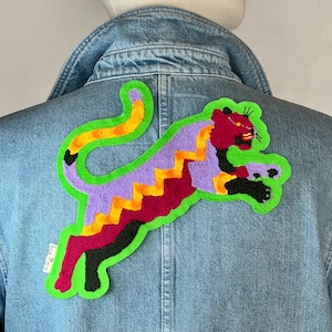 Peut inclure: Veste en jean avec un écusson brodé d'une panthère colorée. L'écusson présente un contour vert, des détails violets, rouges, jaunes et noirs. La panthère saute sur le dos de la veste.