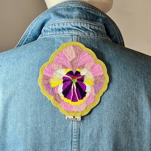 Peut inclure: Une veste en jean bleu clair avec un grand écusson brodé circulaire au dos. L'écusson représente une pensée stylisée dans des tons de rose, violet, jaune et blanc, avec une bordure festonnée en feutre vert olive et une petite étiquette blanche.