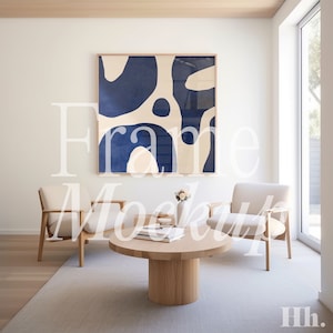 May include: A living room interior design mockup featuring a framed abstract art print with blue and cream colours. Two beige armchairs are positioned either side of a wooden coffee table with books on top. The room has a light wood floor and a large window.