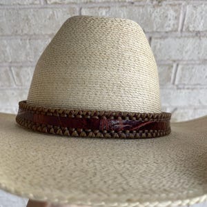May include: A straw cowboy hat with a brown leather band. The hat has a wide brim and a tall crown. The leather band is intricately stitched and adds a touch of rustic elegance.