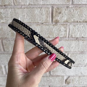 May include: A black and beige snake skin pattern choker necklace. The choker is held in a hand with pink nail polish. The choker is made of a snake skin pattern with black braided edges.