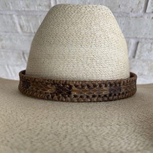 May include: A straw-colored cowboy hat with a brown snakeskin band. The hat has a wide brim and a tall, rounded crown. The snakeskin band is intricately patterned with shades of brown and tan.