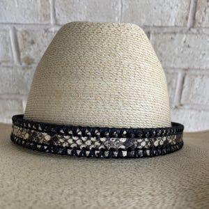 May include: A natural straw hat with a black and snakeskin patterned band. The hat has a wide brim and a rounded crown. The band is made of a woven material and is decorated with a snakeskin pattern.
