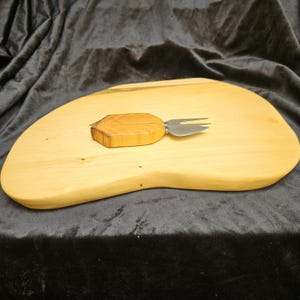 Könnte beinhalten: Ein helles Käsebrett aus Holz mit einer einzigartigen, organischen Form. Eine Käsegabel mit Holzgriff und Edelstahlgabel liegt auf dem Brett. Das Brett ist aus hellem Holz gefertigt.