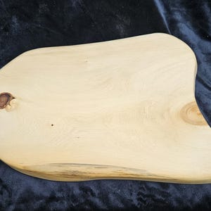 May include: A light-coloured wooden serving board with an irregular, organic shape and a smooth, sanded surface. The board features two distinct natural wood knots, one dark and circular on the left and one lighter, smaller knot on the right side.