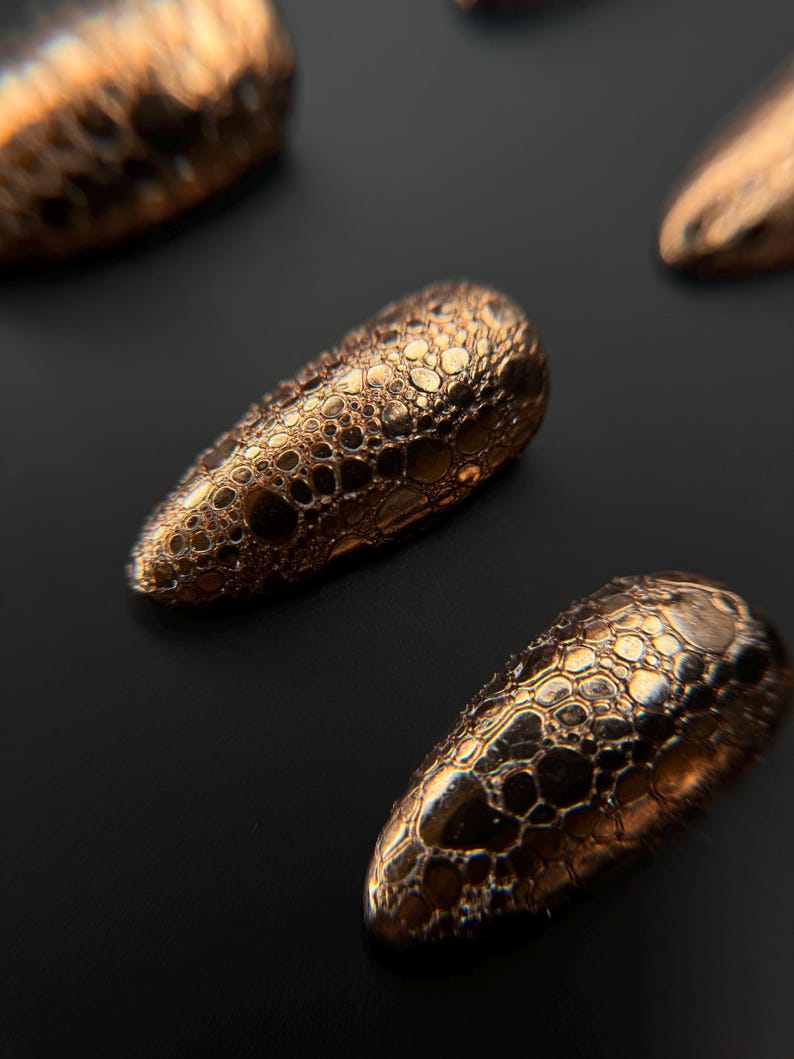 May include: Close-up of several bronze-colored, textured nail tips arranged on a dark surface. The tips have a bubbly, honeycomb-like pattern, reflecting light and creating a metallic sheen. The overall effect is one of luxury and intricate design.