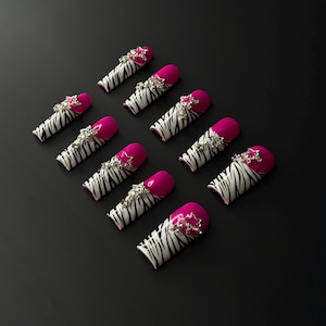 May include: A set of ten artificial nails with a zebra print design in black and white. The tips are painted a bright pink, and each nail is adorned with a silver star-shaped jewel. The nails are arranged on a black surface.