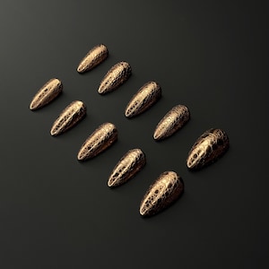 May include: Ten gold-colored, textured nail tips arranged on a black surface. The nail tips have a pointed shape and a scale-like pattern. The overall aesthetic is elegant and sophisticated, suitable for nail art.