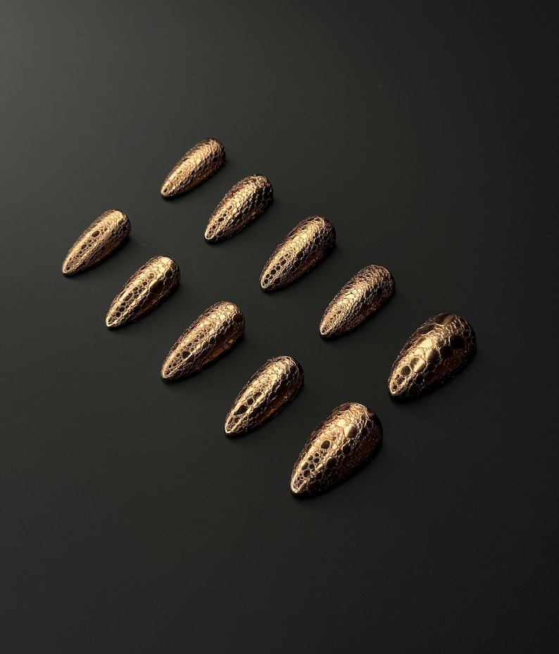 May include: Ten gold-colored, textured nail tips arranged on a black surface. The nail tips have a pointed shape and a scale-like pattern. The overall aesthetic is elegant and sophisticated, suitable for nail art.