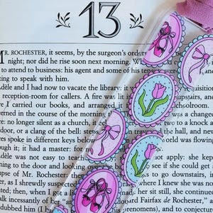 May include: A close-up of a book with a pink and blue bookmark featuring a tulip, ballet shoes, and a bow. The bookmark is on top of the page with the number "13" printed on it. The text on the page reads "Mr. Rochester, it seems, by the surgeon's orders..."