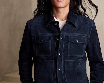 Può includere: Una giacca in camoscio blu navy scuro con colletto classico, chiusura frontale con bottoni metallici e due tasche sul petto con patta abbottonata. La giacca è indossata sopra una maglia bianca semplice, con un design strutturato.