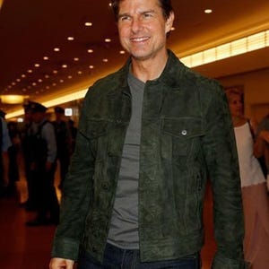 Upper-body view of a man smiling in an olive green suede jacket and grey t-shirt in a brightly lit hallway.