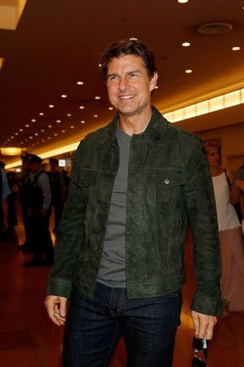Upper-body view of a man smiling in an olive green suede jacket and grey t-shirt in a brightly lit hallway.