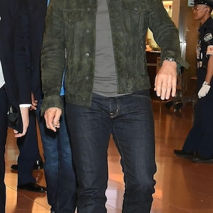 Full-length shot of a man walking while wearing dark sunglasses, an olive green suede jacket, dark jeans, and black boots.