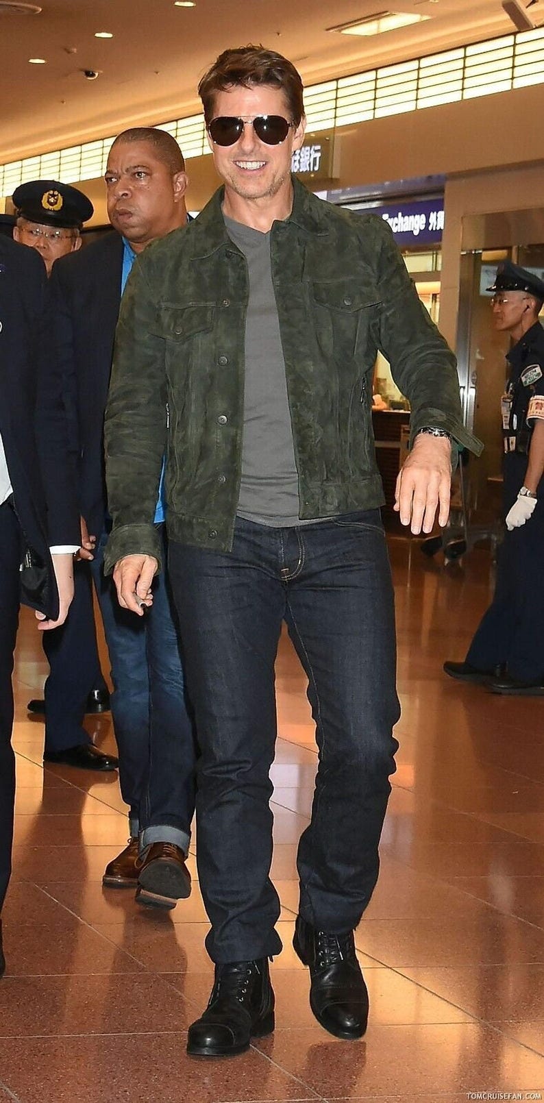 Full-length shot of a man walking while wearing dark sunglasses, an olive green suede jacket, dark jeans, and black boots.
