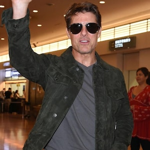 A man smiling and waving with one hand while wearing dark sunglasses and a dark green suede jacket.