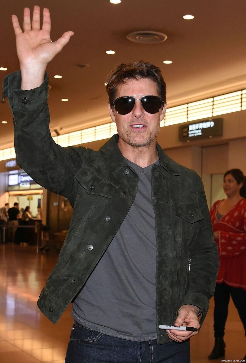 A man smiling and waving with one hand while wearing dark sunglasses and a dark green suede jacket.