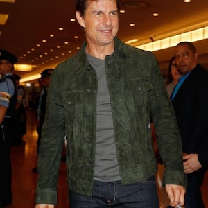 A man wearing an olive green suede jacket, grey t-shirt, and dark jeans indoors in a public setting.