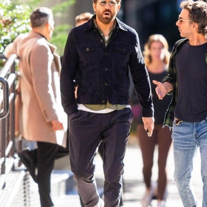 Puede incluir: Un hombre con una chaqueta vaquera azul oscuro, una camiseta verde, pantalones azul oscuro y zapatillas blancas. Camina por la calle y habla con otro hombre.
