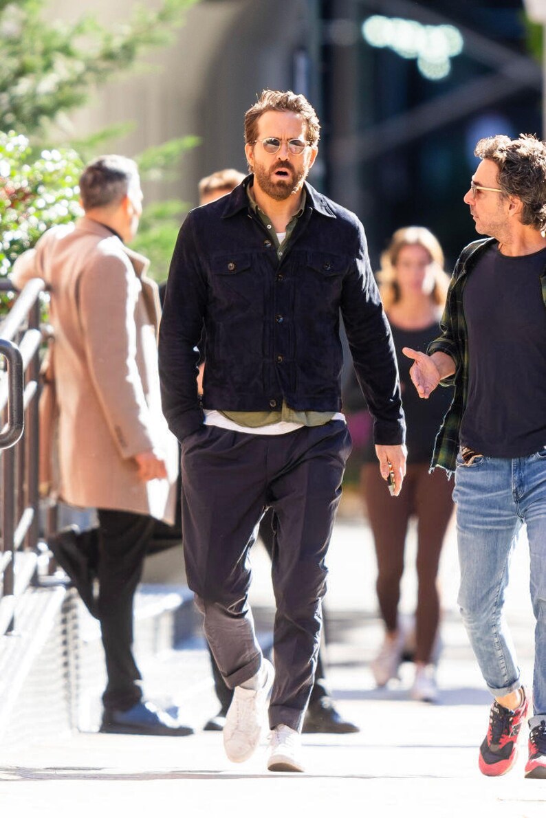 Puede incluir: Un hombre con una chaqueta vaquera azul oscuro, una camiseta verde, pantalones azul oscuro y zapatillas blancas. Camina por la calle y habla con otro hombre.