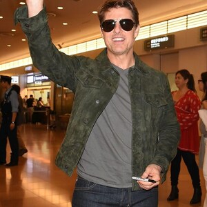 Medium shot of a smiling man wearing dark sunglasses, an olive green suede jacket, and dark jeans while raising a hand to wave.