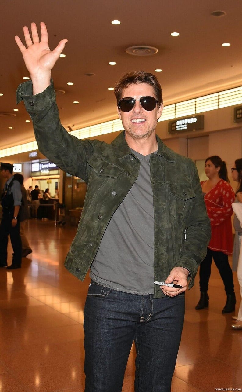 Medium shot of a smiling man wearing dark sunglasses, an olive green suede jacket, and dark jeans while raising a hand to wave.