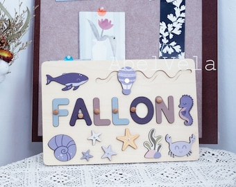 May include: A wooden personalized name puzzle with the name "FALLON" in purple, blue, and brown letters. The puzzle features a variety of sea-themed illustrations, including a whale, a hot air balloon, a starfish, a seashell, a crab, and a seahorse.
