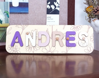 May include: A wooden name puzzle with the name "ANDRES" spelled out in colorful letters. The letters are painted in shades of purple, pink, and white, and are set against a background of a floral pattern.