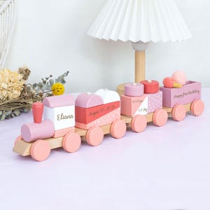 May include: A wooden toy train in shades of pink, white, and coral. The train cars are decorated with geometric patterns and text, including "Eliana" and "August 30, 2025." The last car has the text "Happy first birthday."