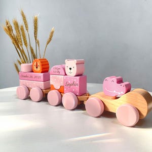 May include: A wooden toy train with pink and orange blocks, featuring a lion, bear, and crocodile. The train has pink wheels and the name "Sophia" is written on one of the blocks. The date "October 26, 2025" is also visible.