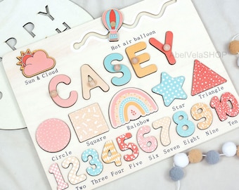 Könnte beinhalten: Holzpuzzle mit dem Namen "CASEY" in bunten Buchstaben, Formen, Zahlen und einem Regenbogen-Design. Enthält einen Heißluftballon und die Wörter "Sun & Cloud".