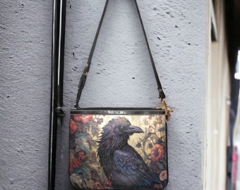 Pode incluir: Uma bolsa de ombro preta com uma ilustração detalhada de um corvo e elementos florais. A bolsa tem uma alça preta e ferragens douradas. A obra de arte apresenta tons de azul, dourado e vermelho.