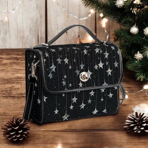 Puede incluir: Bolso de mano negro con estampado de estrellas. El bolso tiene un asa superior y una correa para el hombro desmontable. El cierre de solapa presenta un cierre plateado. El bolso está decorado con estrellas colgantes y pequeños puntos.