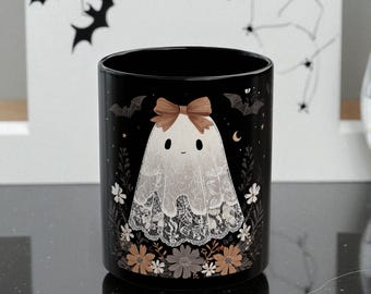 May include: Black ceramic mug featuring a cartoon ghost with a lace veil and a brown bow. The mug is decorated with bats, flowers, and a crescent moon. A Halloween-themed design.